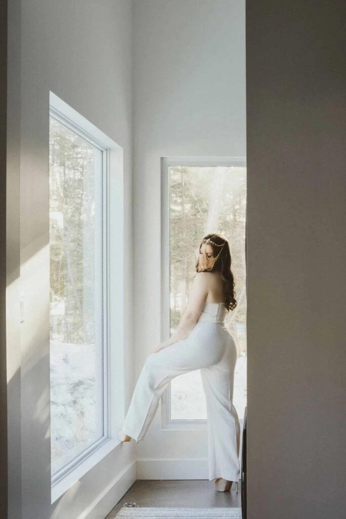 Ava Marie, clad in a white outfit, sits peacefully on a windowsill. She's soaking in the tranquility of her sunlit room while admiring the view of a snow-covered landscape outside. Ava Marie Halifax's Elite Independent Companion
